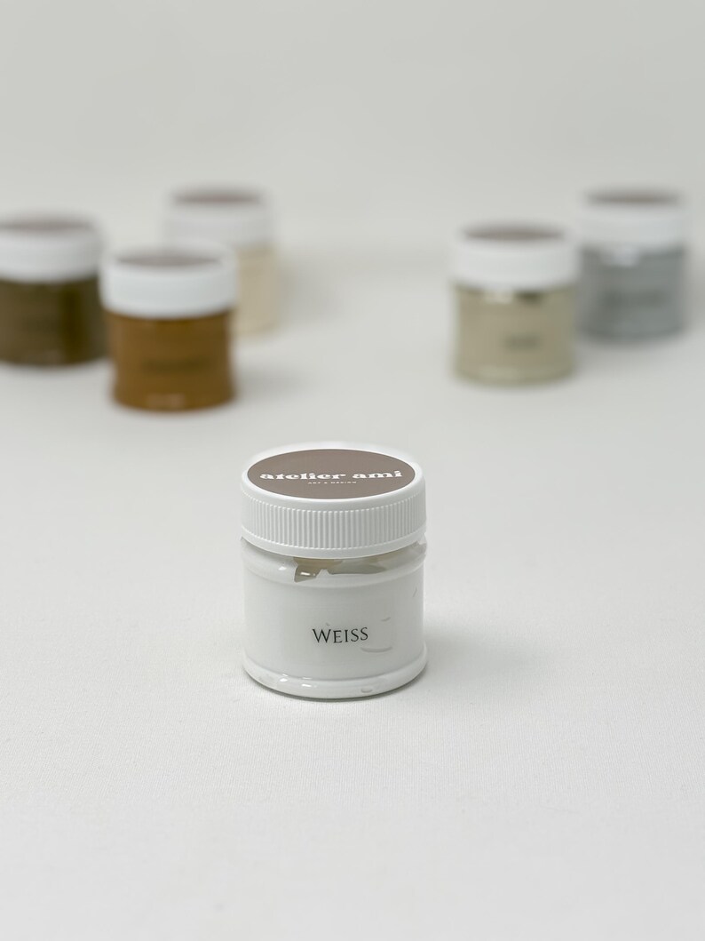 May include: A collection of small, white paint jars with white lids. The front jar has the word "WEISS" printed on it. The lids are brown and have the text "atelier ami" printed on them. Other jars in the background are various shades of brown and gray.