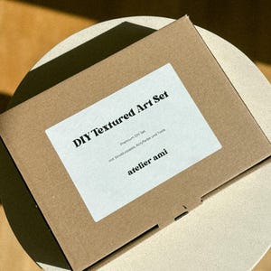 May include: A brown cardboard box labeled "DIY Textured Art Set" with a white label. The label includes the text "Premium DIY Set" and "atelier ami". The box is on a light-colored surface.