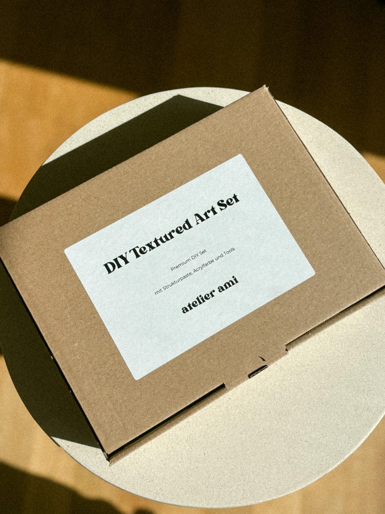 May include: A brown cardboard box labeled "DIY Textured Art Set" with a white label. The label includes the text "Premium DIY Set" and "atelier ami". The box is on a light-colored surface.