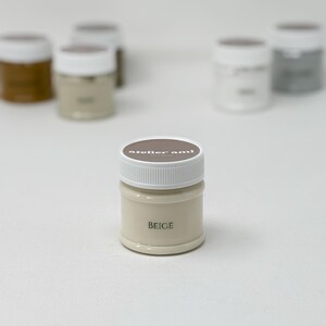 May include: A small, beige-colored paint jar with a white lid. The word "BEIGE" is printed on the front of the jar. The jar is part of a collection of paint jars, some of which are visible in the background.