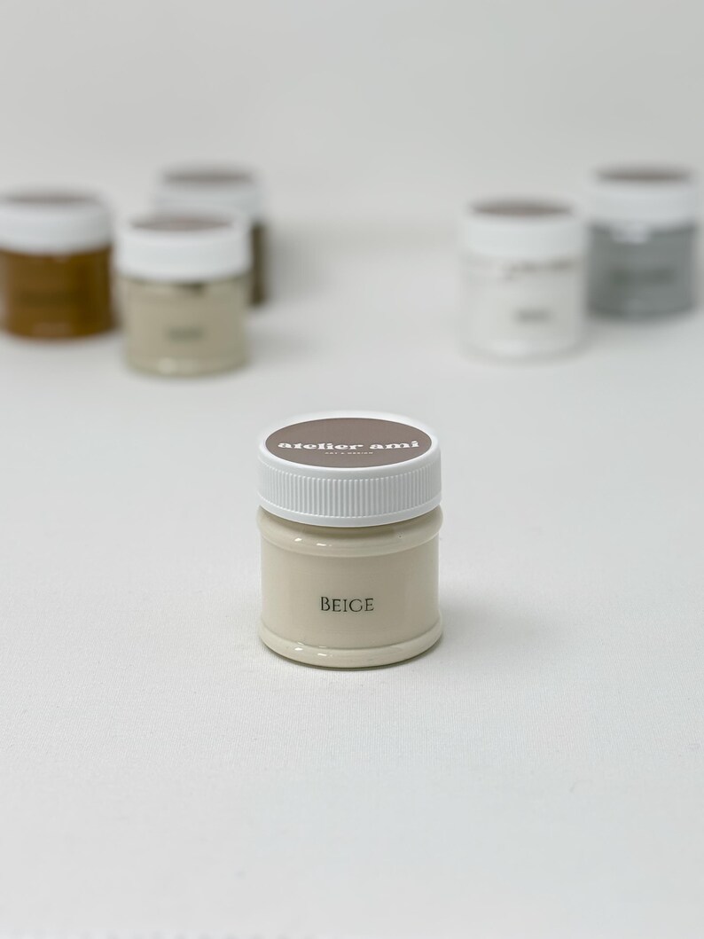 May include: A small, beige-colored paint jar with a white lid. The word "BEIGE" is printed on the front of the jar. The jar is part of a collection of paint jars, some of which are visible in the background.