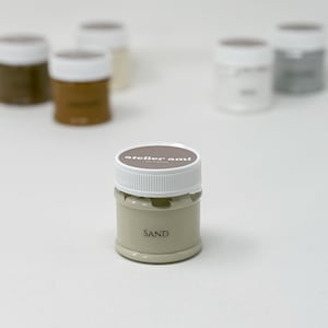 May include: A collection of small paint jars with white lids. The front jar is labeled "SAND" in a neutral beige color. The lids have the text "atelier ami" printed on them. Other jars are in shades of brown, beige, and gray.