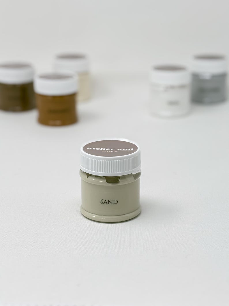 May include: A collection of small paint jars with white lids. The front jar is labeled "SAND" in a neutral beige color. The lids have the text "atelier ami" printed on them. Other jars are in shades of brown, beige, and gray.