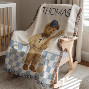 May include: Cream-colored throw blanket with a baseball-themed teddy bear illustration. The blanket features the name "THOMAS" in navy blue letters. The bear wears a baseball cap and uniform, holding a bat. The bottom has a blue and white checkered pattern.