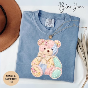 May include: A blue Comfort Colors t-shirt with a patchwork teddy bear design. The bear features floral, gingham, and polka dot patterns in pink, yellow, and mint green. The shirt has the text "Blue Jean" and "Premium Comfort Tee". A gold necklace is draped across the shirt.