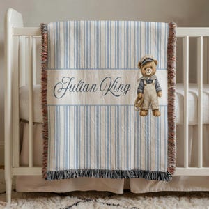 May include: A baby blanket with a blue and white striped pattern and a brown fringe. The blanket features the name "Julian King" in script and a cartoon bear wearing a sailor outfit.