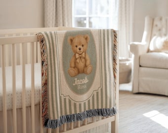 May include: A beige and green striped baby blanket with a teddy bear illustration and the name "Jacob Ross". The blanket is draped over a white crib. The teddy bear has a bow tie.