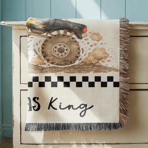 May include: Cream-colored throw blanket featuring a watercolor illustration of a monster truck kicking up dirt. The design includes a black and white checkered pattern and the text "IS King". The blanket has fringed edges.