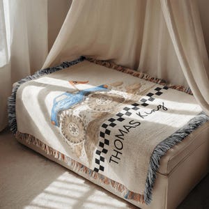 Op de afbeelding: Een beige sprei met een blauw tractorontwerp en de naam "Thomas King" in zwart schrift. De sprei heeft een geblokte rand en gefranjerde randen. De tractor heeft grote wielen en een kleine oranje vlag.