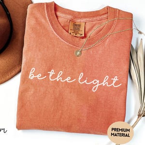 May include: A coral-colored t-shirt with the phrase "be the light" in white script. A gold necklace with a pendant rests on the shirt. A brown hat and a dried palm leaf are also in the image. The shirt has a "Premium Material" label.
