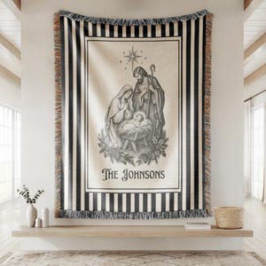 May include: A woven throw blanket featuring a black and white nativity scene with Mary, Joseph, and the infant Jesus under a star. The blanket has a black and white striped border with fringe and the text "The Johnsons" printed at the bottom.