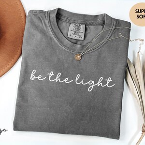 May include: A gray t-shirt with the phrase "be the light" in white script. The shirt has a crew neck and short sleeves. A gold pendant necklace is draped across the shirt. A brown hat and dried plant are in the background.
