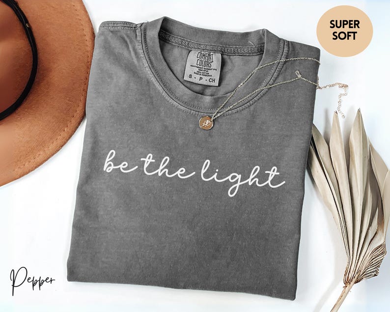 May include: A gray t-shirt with the phrase "be the light" in white script. The shirt has a crew neck and short sleeves. A gold pendant necklace is draped across the shirt. A brown hat and dried plant are in the background.