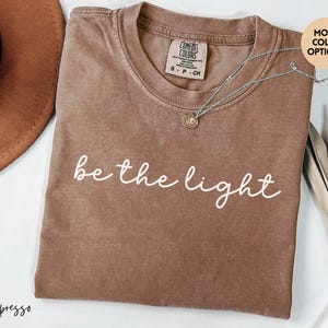 May include: Espresso-colored t-shirt with the phrase "be the light" in white script. The shirt has a crew neck and short sleeves. A silver necklace with a small pendant rests on the shirt. The shirt is a Comfort Colors brand.