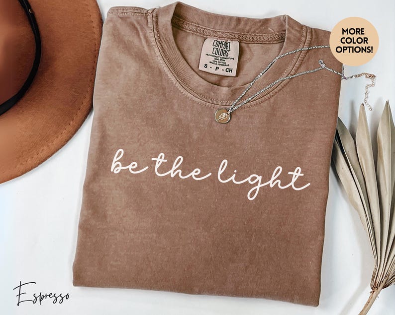 May include: Espresso-colored t-shirt with the phrase "be the light" in white script. The shirt has a crew neck and short sleeves. A silver necklace with a small pendant rests on the shirt. The shirt is a Comfort Colors brand.