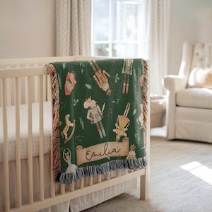 May include: A green woven blanket with a fringe, draped over a crib. The blanket features a repeating pattern of Nutcracker-themed illustrations in various colors. A banner with the name "Emilia" is also visible.
