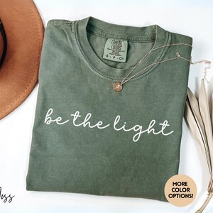 May include: A moss green t-shirt with the words "be the light" in white script. A gold necklace with a pendant rests on the shirt. A brown hat and dried plant stems are also in the image. The shirt has a tag that says "Comfort Colors".