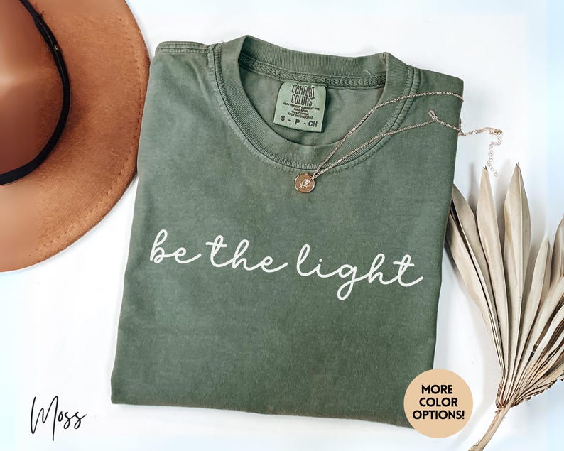 May include: A moss green t-shirt with the words "be the light" in white script. A gold necklace with a pendant rests on the shirt. A brown hat and dried plant stems are also in the image. The shirt has a tag that says "Comfort Colors".