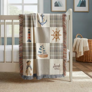 May include: A nautical-themed baby blanket with a patchwork design. The blanket features squares with an anchor, lighthouse, ship's wheel, sailboat, and a teddy bear. The blanket has a plaid pattern and the name "Jacob KING" is printed on the bottom right.