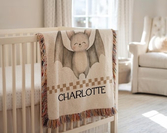 May include: A beige baby blanket with a smiling bat illustration and the name "CHARLOTTE" in black letters. The blanket has a checkered border and colorful fringe. The blanket is draped over a white crib.