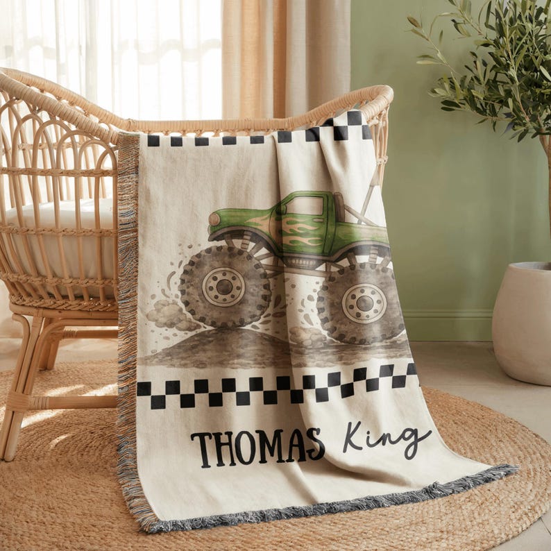 May include: A beige baby blanket with a green monster truck design. The truck has large tires and flames. The blanket has a black and white checkered border and the name "THOMAS King" in black script.