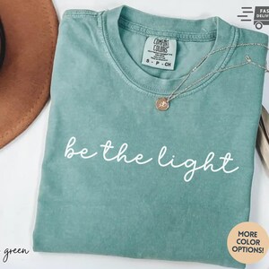 May include: Light green t-shirt with the phrase "be the light" in white script. A gold-colored necklace with a pendant rests on the shirt. The shirt is folded and has a Comfort Colors tag.