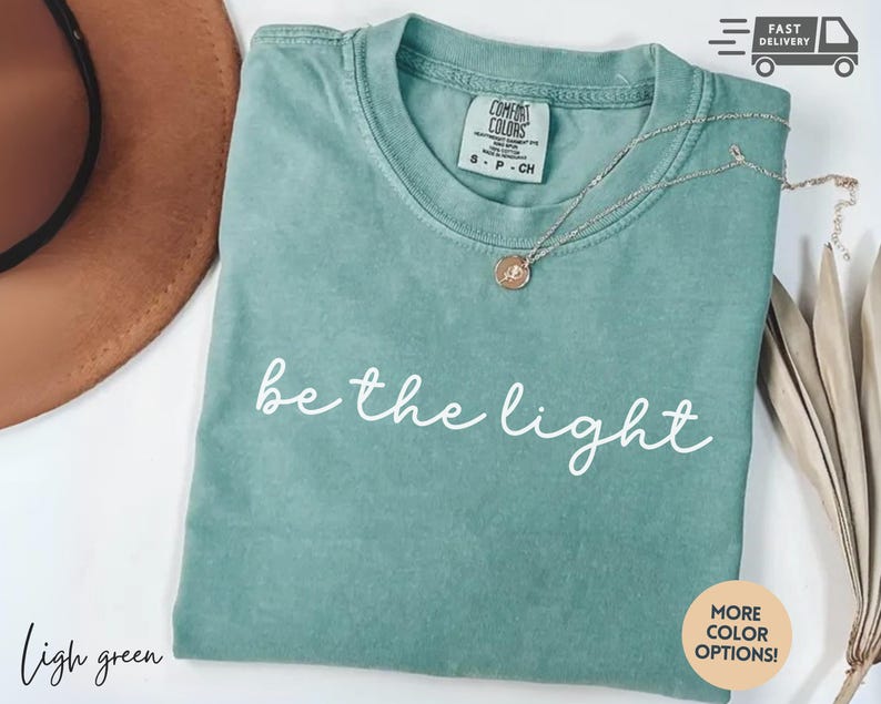 May include: Light green t-shirt with the phrase "be the light" in white script. A gold-colored necklace with a pendant rests on the shirt. The shirt is folded and has a Comfort Colors tag.