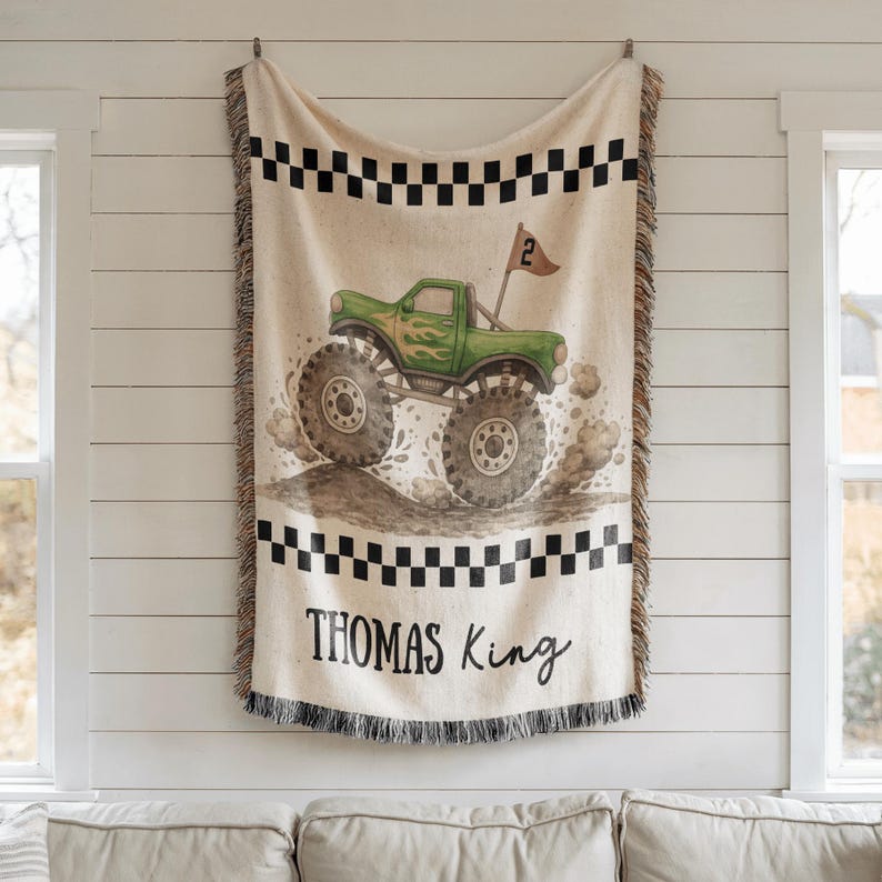 May include: A beige throw blanket featuring a green monster truck design with flames and large tires. The blanket has a black and white checkered border and the name "THOMAS KING" in black script. The blanket has a fringed edge.