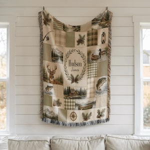 K&ouml;nnte beinhalten: Eine gewebte Wohndecke mit Patchwork-Design und naturbezogenen Illustrationen wie einem Hirsch, Fischen, Enten, einer H&uuml;tte, einer Laterne und einem Kompass. In der Mitte befindet sich ein Lorbeerkranz mit dem Schriftzug Hudson James.