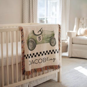 May include: A beige baby blanket with a vintage green race car design. The blanket features the number 5 on the car, a black and white checkered pattern, and the name "JACOB King" in black script. The blanket has a fringed edge.