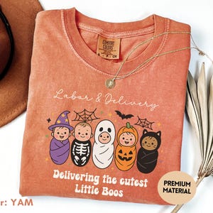 May include: A yam-colored t-shirt with a crew neck and short sleeves. The shirt features a graphic of five babies dressed in Halloween costumes, with the text "Labor & Delivery" and "Delivering the cutest Little Boos."