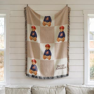 May include: A beige and brown checkered throw blanket with a repeating pattern of teddy bears wearing red caps and blue sweaters with an American flag design. The blanket has a fringe and the name "JACOB JOHNSON" is printed on the bottom right.