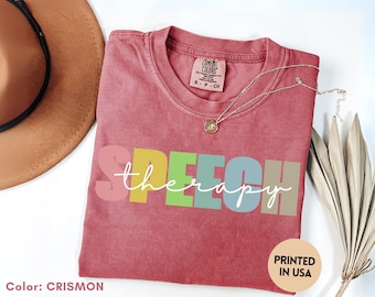 May include: Crimson-colored t-shirt with the words "SPEECH therapy" in colorful block letters. The shirt has a crew neck and short sleeves. A gold necklace with a pendant is draped across the shirt. The words "PRINTED IN USA" are printed on the bottom right.