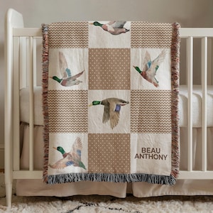May include: A beige and brown patchwork blanket with images of flying ducks. The blanket has a fringed edge and is draped over a crib. The blanket has a repeating pattern of ducks, polka dots, and chevron designs. The name "BEAU ANTHONY" is printed on the blanket.