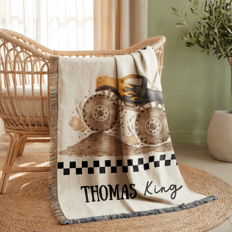 May include: A beige baby blanket with a monster truck design. The truck is yellow and black, with large brown tires, kicking up dirt. The blanket has a black and white checkered border and the name "THOMAS King" in black script.