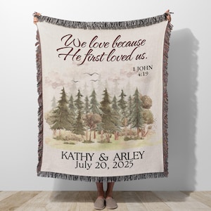 May include: A beige throw blanket with a forest scene and the text "We love because He first loved us." The blanket includes the names "KATHY & ARLEY" and the date "July 20, 2025."