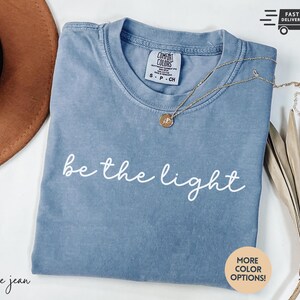 May include: Blue jean colored t-shirt with the phrase "be the light" in white script. A gold necklace with a pendant rests on the shirt. The shirt is folded and displayed with a brown hat and dried plant.
