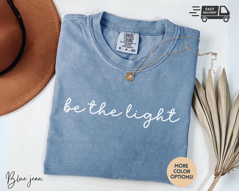 May include: Blue jean colored t-shirt with the phrase "be the light" in white script. A gold necklace with a pendant rests on the shirt. The shirt is folded and displayed with a brown hat and dried plant.