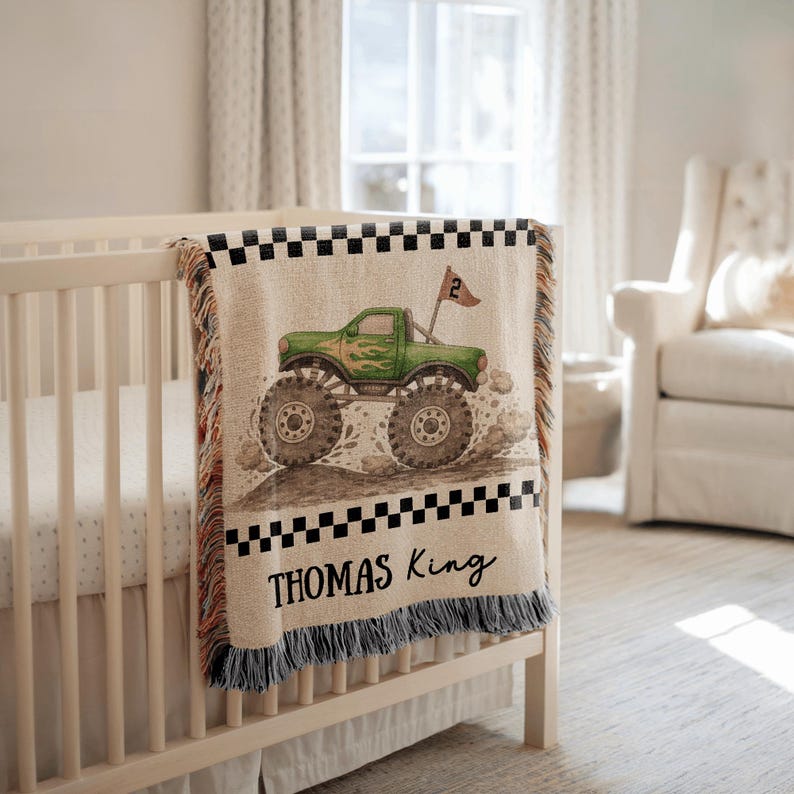 May include: A beige baby blanket with a green monster truck design. The truck has large tires and a flag with the number 2. The blanket has a black and white checkered border and the name "THOMAS King" printed in black.