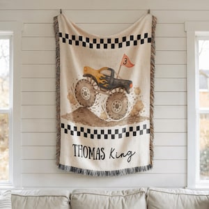 May include: A beige throw blanket featuring a monster truck design with black and yellow flames, a red flag with the number 3, and the name "THOMAS KING". The blanket has a black and white checkered border and fringe.