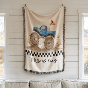 Op de afbeelding: Een beige sprei met een blauw monstertruckontwerp. De truck heeft grote banden en vlammen, met een vlag met de tekst "2". De sprei heeft een zwart-wit geblokte rand en de naam "THOMAS King" staat erop.