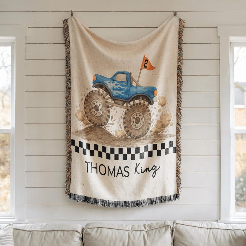 Op de afbeelding: Een beige sprei met een blauw monstertruckontwerp. De truck heeft grote banden en vlammen, met een vlag met de tekst "2". De sprei heeft een zwart-wit geblokte rand en de naam "THOMAS King" staat erop.