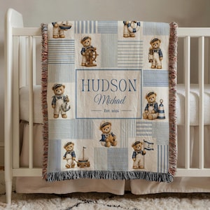 May include: A woven throw blanket featuring a nautical theme with teddy bears in sailor outfits, blue and white stripes, and a central rectangular panel displaying the text HUDSON Michael EST. 2026 with fringed edges on the top and bottom.