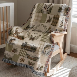 K&ouml;nnte beinhalten: Eine gewebte Wohndecke mit Patchwork-Design und Outdoor-Motiven wie Hirsch, Fischen, H&uuml;tte, Kanu, Tannenzapfen und Laterne. In der Mitte befindet sich ein Kranz-Motiv mit dem Schriftzug Hudson James in einer geschwungenen Schreibschrift.
