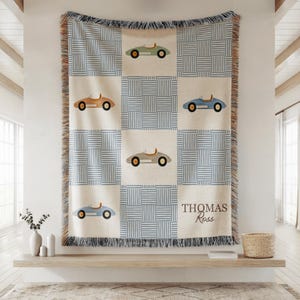 May include: Woven throw blanket with a repeating pattern of blue and white squares and vintage car illustrations in various colors. The blanket has a fringed edge and the name "Thomas Ross" is displayed in a corner.