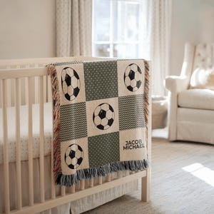 May include: A personalized baby blanket with a soccer ball design. The blanket features a patchwork pattern with soccer balls, geometric shapes, and the name "JACOB MICHAEL" in a square. The blanket is draped over a crib.