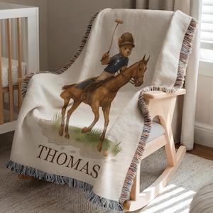 May include: Cream-colored throw blanket draped over a wooden rocking chair. The blanket features a watercolor illustration of a teddy bear playing polo on a brown horse. The name "THOMAS" is printed on the blanket.