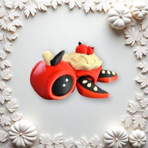 May include: A red and black turtle figurine with a cream-colored shell and red accents. The turtle is carrying a small red and cream-colored pumpkin on its back.