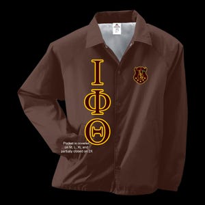 Puede incluir: Una chaqueta marrón con un diseño de letras griegas doradas y granate. Las letras deletrean "IΦΘ". La chaqueta tiene un bolsillo en el lado izquierdo y un cierre de botón.