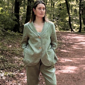 K&ouml;nnte beinhalten: Ein hellgr&uuml;ner Leinenanzug, bestehend aus einem Blazer mit zwei Kn&ouml;pfen und einer passenden Hose. Der Anzug wird mit einem hellblauen Oberteil und dunkelblauen Slip-On-Schuhen getragen. Der Hintergrund ist ein Waldweg.